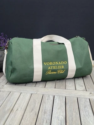 Voronado Riviera Duffelbag - Stilren grön shoppingväska från Voronado Atelier med vita detaljer och axelrem. Väskan har broderad gul text på framsidan och är tillverkad i slitstark canvas. Perfekt för att bära med sig allt man behöver till skolan, gymmet eller stan.