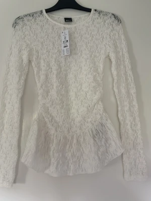 Vit spetsblus med volang - Säljer en superfin vit spetsblus med lång ärm och volang nedtill. Blusen har en figurnära passform och är helt täckt i spets med en glimt av blommor. Perfekt att styla med jeans eller kjol för en trendig look. ny pris 300kr, nu 200. aldrig använd. prislapp kvar❣️