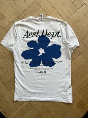 Vit t-shirt med blått tryck Aest Dept. - Snygg vit t-shirt med stor blå blomma och texten 'Aest. Dept.' samt 'ADAPTIVE REALITY' på ryggen. Framtill finns en liten logga på bröstet. T-shirten har rund hals och korta ärmar, perfekt för en clean och modern stil.