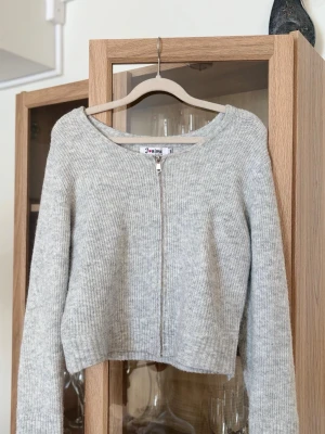 Ljusgrå stickad zip-kofta - Ljusgrå stickad kofta med dragkedja framtill och rund halsringning. Koftan har långa ärmar och är något croppad i modellen. Liten i storleken