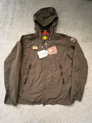 Grön vindjacka från Parajumpers - Snygg brun vindjacka med huva och dragkedja framtill. Jackan har två framfickor med dragkedja, patchar på bröstet och ärmen samt justerbar nederkant. Tillverkad i lätt och slitstarkt material, perfekt för blåsiga dagar.