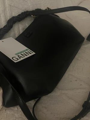 Svart handväska från Ganni - Stilren svart handväska från Ganni i slätt material med flätat handtag och justerbar axelrem.