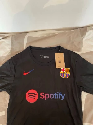 Svart FC Barcelona matchtröja Nike - Snygg svart FC Barcelona matchtröja från Nike med klubbmärke på bröstet och röd Nike-logga. Spotify-logga i rött och lila framtill. Diskreta Barcelona-emblem i tyget. Perfekt för dig som älskar fotboll och vill ha en stilren supportertröja.