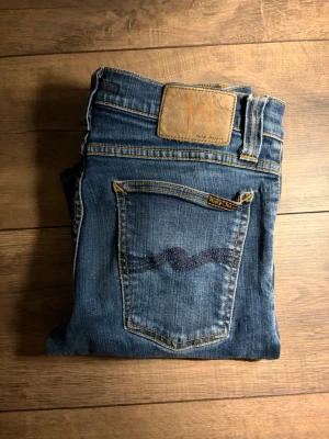 Nudie jeans  - Säljer ett par klassiska blå jeans från Nudie Jeans med snyggt sliten look och ikonisk bakficka med vågig söm. Jeansen har normal midja, smal passform och är gjorda i mjukt denimtyg. Perfekta till din vardagsstil!