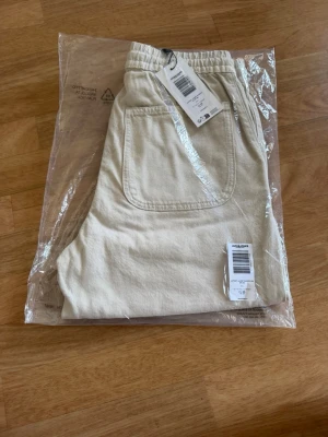 Beige shorts med snörning - Säljer ett par beige shorts med elastisk midja och snörning framtill. Shortsen har stora fickor både fram och bak och är tillverkade i ett mjukt bomullsmaterial. Perfekta för varma dagar och enkel att matcha med olika stilar. 