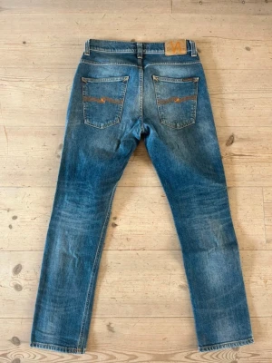  Nudie Jeans - Storleken är 31/30 Säljer ett par klassiska blå jeans från Nudie Jeans med raka ben och snygg slitning framtill. Jeansen har fem fickor, kontrastsömmar och Nudie-logga på bakfickan. Perfekta för en avslappnad och trendig look.