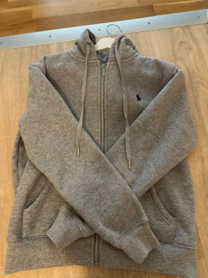Grå hoodie från Polo Ralph Lauren - Säljer en grå hoodie från Polo Ralph Lauren med dragkedja, huva och dragsnören. Hoodien har fickor på sidorna, ribbade muddar och ett mörkblått broderat logotyp på bröstet. Insidan är fodrad med mjuk teddy för extra komfort.