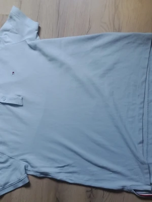 Tommy Hilfiger piké - Snygg ljusblå Tommy Hilfiger piké med korta ärmar. Enkel och stilren design som passar till det mesta. Det finns en svart fläck (bild 2) men annars är den riktigt bra skick! Använd fåtal gånger. Ps Pris kan diskuteras!