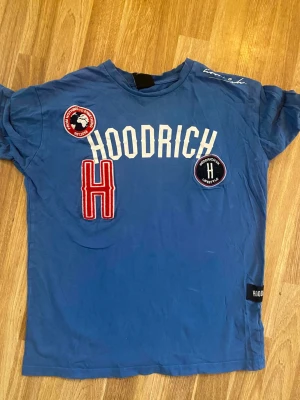  Hoodrich t-shirt - Blå t-shirt från Hoodrich med vita tryck och flera patches på bröstet, inklusive ett stort rött H och två runda märken. T-shirten har rund hals och korta ärmar. Perfekt för dig som gillar streetwear och vill sticka ut.