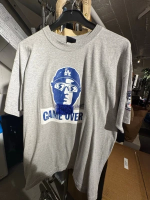Grå t-shirt med LA-tryck - Säljer en grå t-shirt med ett stort blått tryck framtill av en person med solglasögon och LA-keps samt texten 'GAME OVER'. T-shirten har korta ärmar och rund hals. Perfekt för dig som gillar sportiga och avslappnade plagg. Finns att hämta Gröndal. 