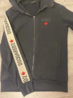 Grå hoodie från Dsquared2 med dragkedja - Snygg grå hoodie från Dsquared2 med dragkedja framtill, huva och fickor. På bröstet finns DSQ2-logga och ett rött lönnlöv, samt en beige detalj med DSQUARED2-tryck och lönnlöv längs ena ärmen. Perfekt för en avslappnad streetwear-look.   Fråga för mer bilder och info