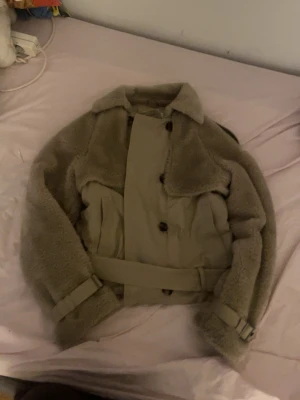 Beige teddyjacka med bälte - Säljer en beige teddyjacka med mjuka, lurviga ärmar och krage. Jackan har en snygg bälte i midjan och detaljerade spännen vid ärmsluten. Stängs med knappar framtill och har en cool, oversized look. Perfekt för dig som vill ha en trendig och mysig jacka.