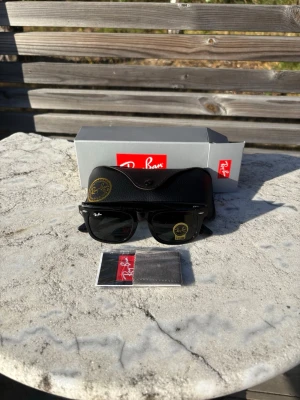 Svarta Ray-Ban Wayfarer solglasögon - Klassiska svarta Ray-Ban Wayfarer solglasögon med tidlös design och mörka glas. Bågarna är i blank svart plast och har Ray-Ban-loggan på både glas och skalmar. Perfekta för soliga dagar och kommer med originalfodral och putsduk.