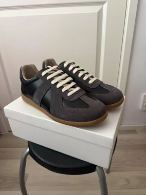Maison Margiela Svarta och gråa 43 - Stilrena sneakers i grått med inslag av svart läder och mockadetaljer. Skorna har vita snören och en klassisk gummisula i beige. Perfekta för en trendig och avslappnad look.