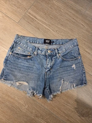 Blå jeansshorts med fransig kant - Snygga blå jeansshorts med fransig och ruffad kant nertill. Klassisk femficksmodell med knapp och dragkedja framtill. Perfekta för sommaren och ger en avslappnad vibe.