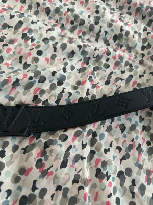 Svart bälte med präglad logga - Snyggt svart bälte i skinn med präglade loggor och detaljer längs hela bandet. Bältet har en stilren design och bred form som gör det till ett statement-piece i garderoben. Perfekt för att lyfta vilken outfit som helst.