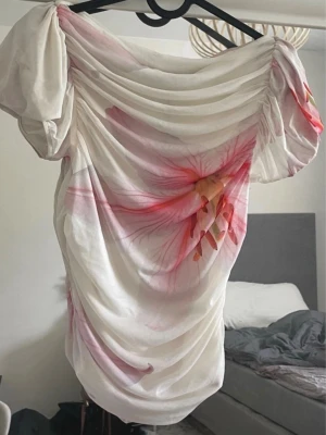 Sommrig topp - Drömmig vit blus med stora rosa blommor och draperad omlottdesign. Toppen har offshoulder-modell, puffiga korta ärmar och ett skirt, flowy tyg som ger en romantisk vibe. Perfekt för dig som gillar feminina detaljer. Har aldrig blivit använd! Har små sminkfläckar på framsidan!!