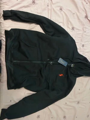 Svart hoodie med dragkedja från Polo - Svart hoodie med dragkedja från Polo Ralph Lauren. Klassisk design med huva, två fickor framtill och den ikoniska röda logotypen broderad på bröstet. Tillverkad i mjukt material som passar perfekt för en avslappnad stil.