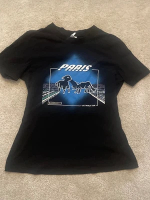 Svart Paris World Tour t-shirt - Säljer en svart t-shirt med tryck fram som visar Paris och två figurer, samt texten 'WORLD TOUR' på ryggen. T-shirten har korta ärmar och en klassisk passform. Perfekt för dig som gillar coola bandtröjor och streetwear.