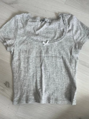 Ljusgrå ribbad topp med spets - Supersöt ljusgrå ribbad topp med korta ärmar, spetskantad urringning och liten rosett framtill. Perfekt för en chill och söt look. Mjuk och skön i bomullsmix, lätt att matcha med jeans eller kjol.
