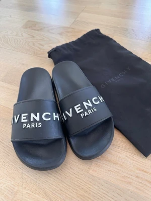Svarta slides från Givenchy - Säljer ett par svarta slides från Givenchy med vit logga på ovandelen. Sandalerna har en klassisk slip-on design och är tillverkade i syntetmaterial. Perfekta för sommaren eller att glida runt i hemma. Medföljer original dustbag.