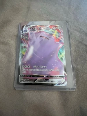 Pokémonkort Ditto VMAX japansk - Säljer ett japanskt Pokémonkort med Ditto VMAX-motiv. Kortet har en holografisk yta och färgstark design med Ditto i lila och regnbågsfärgad bakgrund. Kortet ligger i ett genomskinligt plastskydd. Perfekt för samlare eller Pokémonfans.