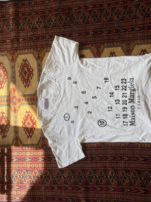 Maison Margiela vit t-shirt med tryck - Vit t-shirt från Maison Margiela med ikoniskt svart siffertryck framtill. Klassisk passform och rund halsringning. Perfekt för dig som gillar minimalistisk och stilren design. Tillverkad i mjuk bomull för skön känsla. 65cm lång