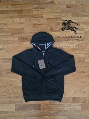 Svart Burberry Hoodie - Letar du efter en stilren och lyxig hoodie? Denna helt nya grå Burberry-hoodie kombinerar elegans med hög komfort. Den har Burberrys ikoniska rutmönster i fodret och en diskret logga på bröstet för en exklusiv touch. Tillverkad av mjukt, slitstarkt material och utrustad med dragkedja samt huva – både praktisk och stilfull.  📌 Skick: Ny med taggar. 📦 Snabb leverans – skickas samma dag (om ej hinder)