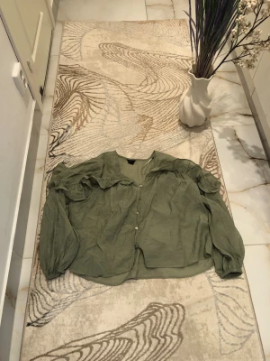 Olivgrön blus med volangärmar - Snygg olivgrön blus i ett tunt, luftigt tyg. Blusen har långa ärmar med volangdetaljer på axlarna och knäppning framtill. Perfekt för dig som gillar en romantisk och trendig stil. Materialet ser ut att vara bomull eller linne.