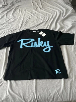 Svart t-shirt med blå Risky-tryck - Snygg svart t-shirt med stort blått 'Risky'-tryck på bröstet och en liten blå logga nere vid fållen. Klassisk passform och korta ärmar, perfekt till jeans eller shorts. Tillverkad i mjuk bomull för skön känsla hela dagen. Oanvänd. 