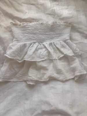 Vit volangkjol  - Supersöt vit kjol med bred smock i midjan och dubbla volanger. Kjolen är kort och har en luftig känsla, perfekt för varma dagar. Tillverkad i ett tunt och mjukt material som ger en flowy look.