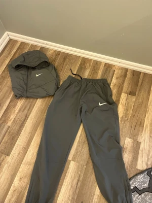 Grått Nike träningsset med huva - Säljer ett grått träningsset från Nike med matchande byxor och jacka. Jackan har huva, dragkedja och två fickor framtill samt vit Nike-logga på bröstet. Byxorna har resår i midjan och vit Nike-logga på benet. Perfekt för träning eller chill. Ett jättte bra kap om du frågar mig för detta priset. Storlekx S priser kan diskuteras vid snabb äffär