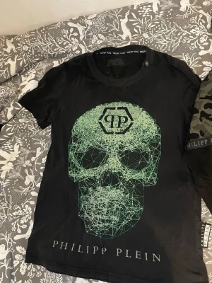 Svart Philipp Plein t-shirt med dödskalle - Snygg svart t-shirt från Philipp Plein med stort grönt dödskalletryck framtill och logga både fram och bak. T-shirten har rund hals och är tillverkad i mjukt material som känns skönt mot huden. Perfekt för dig som gillar edgy streetwear.