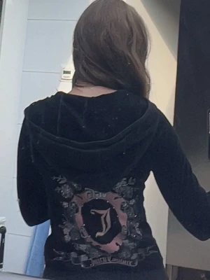 Svart velour hoodie kofta Juicy Couture - Säljer en svart velour kofta från Juicy Couture med dragkedja framtill. Stor broderad logga och emblem i rosa och silver på ryggen samt mindre logga på bröstet. Hoodien har huva och fickor framtill. Perfekt för en avslappnad och trendig look. Sitter tajt. 