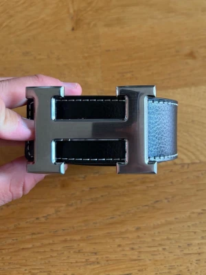 Svart skärp med stor H-spänne - Snyggt svart bälte i skinn med ett stort, silverfärgat H-format spänne. Bältet har vita sömmar längs kanterna som ger en schysst kontrast. Perfekt accessoar för att lyfta vilken outfit som helst.