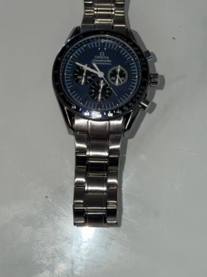 Omega Speedmaster silverklocka - Stilren Omega Speedmaster med klassisk rund urtavla i svart och detaljer i vitt. Klockan har en robust länkarmband i silverfärgat metall och flera små urtavlor för extra funktioner. Perfekt för dig som gillar tidlös design och kvalitet.