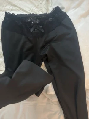 Svarta leggings med spets och rosett - Säljer ett par svarta leggings med bred spetskant och liten rosett framtill i midjan. Leggingsen har en mjuk och stretchig passform.