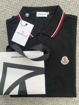 Svart pikétröja från Moncler - Snygg svart pikétröja från Moncler med klassisk krage och knappar framtill. Kragen har röda och vita ränder som ger en sportig touch. Moncler-logga är broderad på bröstet. Perfekt till en stilren och avslappnad look.