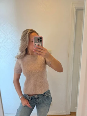 Beige stickad topp med kort ärm - Mysig beige stickad topp med korta ärmar och rund hals. Toppen har en enkel och stilren design som passar perfekt till jeans eller kjol. Materialet ser fluffigt och mjukt ut, vilket ger en avslappnad men ändå trendig look.