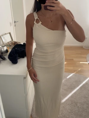 Beige oneshoulder långklänning - Säljer en beige oneshoulder långklänning med snygg knyt- och ringdetalj på axeln. Klänningen har en figurnära passform och är tillverkad i ett mjukt, stretchigt material som ger en smickrande siluett. Perfekt för dig som vill ha en stilren och trendig look.