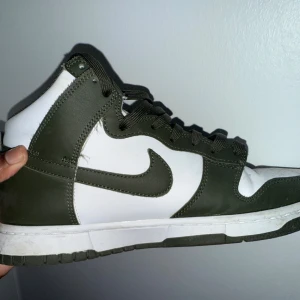 Nike Dunk High olivgrön/vit sneaker - Snygga Nike Dunk High sneakers i olivgrönt och vitt. Skorna har klassisk hög siluett, vit tåbox och sidor, samt olivgröna detaljer och snörning. Ikoniska swoosh-loggan på sidan och slitstark gummisula. Perfekta för dig som gillar streetwear och sportig stil.