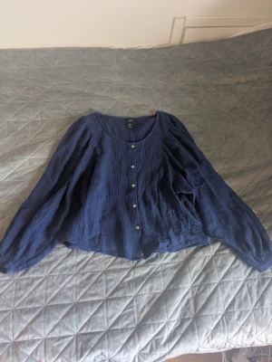 Blå långärmad blus från Lindex - Säljer en blå långärmad blus från Lindex med knappar framtill. Blusen har en rund halsringning och är gjord i ett mjukt, krinklat bomullstyg. 