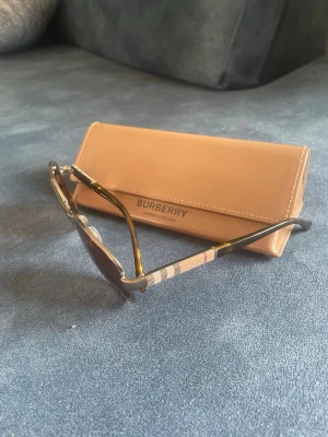 Randiga solglasögon från Burberry - Snygga solglasögon från Burberry med klassiskt randigt mönster på skalmarna i beige, svart och brunt. Glasögonen har en stilren form och kommer med ett beige fodral med Burberry-logga. Perfekta för att lyfta din look under soliga dagar.
