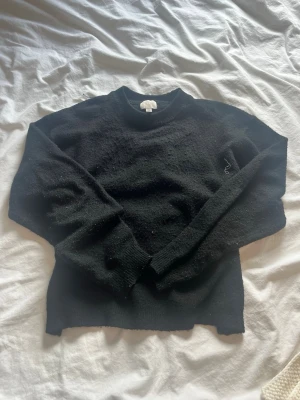 Svart stickad tröja med rund hals - En enkel svart stickad tröja med rund hals och långa ärmar. Tröjan har ribbstickade muddar vid ärmslut och nederkant samt en diskret broderad detalj på bröstet. Perfekt för lager på lager under kyliga dagar.