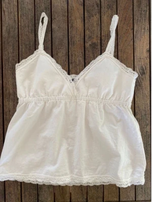 Brandy Melville vit babydolltop med spets - Söt vit topp (”Edith Lace Tank”) från Brandy som inte längre säljs på hemsidan. Den har spetskanter, v-ringning och en rynkad midja vilket skapar en snygg silhuett. Endast testad och säljer då den inte kommer till användning. Gjord i 100% bomull. Passar XS och S. Pris går att diskutera🩷