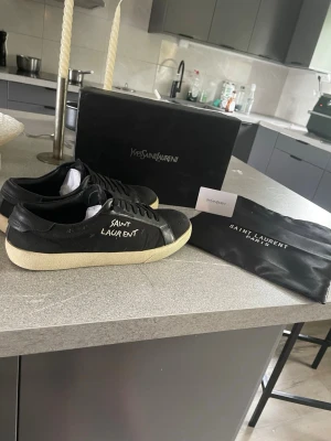 Svarta Saint Laurent sneakers - Svarta sneakers från Saint Laurent med vit sula och handskriven logga på sidan. Skorna har klassisk låg profil, rund tå och snörning framtill. Tillverkade i läder med detaljer i textil. Kommer med originalkartong och dustbag. På bild 4 kan man se att limmet från skon har gått upp eller nåt men det blir bara en öppning när man drar ut den och inget som påverkar skon