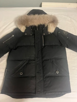 Moose knuckles Svart dunjacka med pälskrage - Snygg svart dunjacka med stor luva och fluffig pälskant. Jackan har flera fickor med dragkedja och knappstängning samt en patch på ärmen. Perfekt för kalla vinterdagar och riktigt varm. Klassisk, rak modell med detaljer i metall.