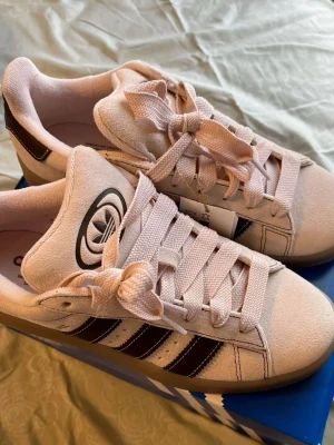 Adidas Campus ljus rosa - Snygga Adidas Campus sneakers i ljus rosa färg med bruna detaljer och klassiska tre ränder på sidan. Skorna har en låg profil, platt sula och Adidas-logga. Perfekta för en clean och stilren look.