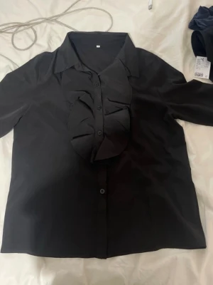 Svart blus med volangdetalj - Stilren svart blus med långa ärmar och volangdetalj längs knappraden framtill. Klassisk krage och knäppning hela vägen ner. Materialet ser ut att vara mjukt och lätt.