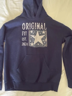 Mörkblå hoodie med stjärntryck - Säljer en mörkblå hoodie med huva och magficka. På bröstet står det 'ORIGINAL FIT EST. 2024' och ett stort stjärntryck med mindre stjärnor runt. Hoodien har ribbade muddar vid ärmslut och nederkant. Perfekt för en avslappnad streetstyle.